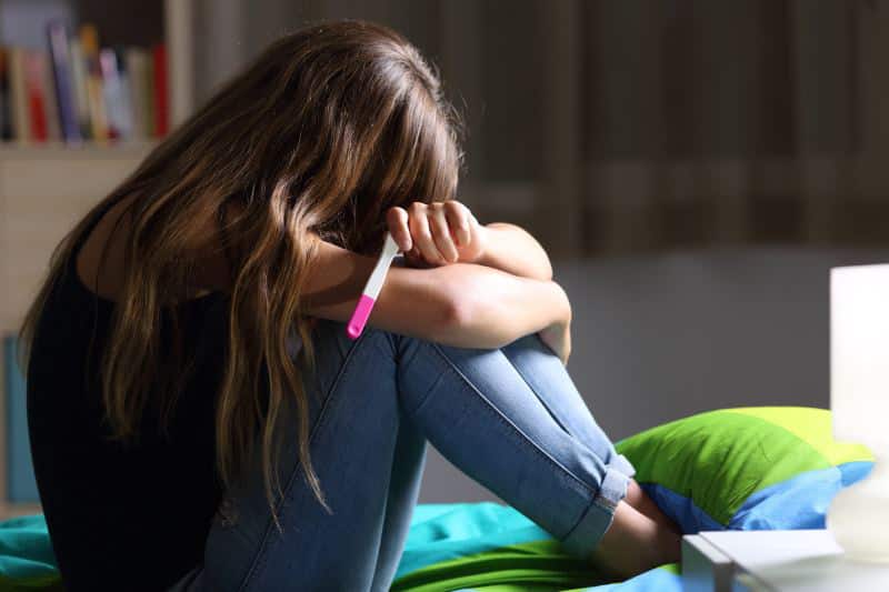 Junges Mädchen sitzt weinend mit einem Schwangerschaftstest auf dem Bett